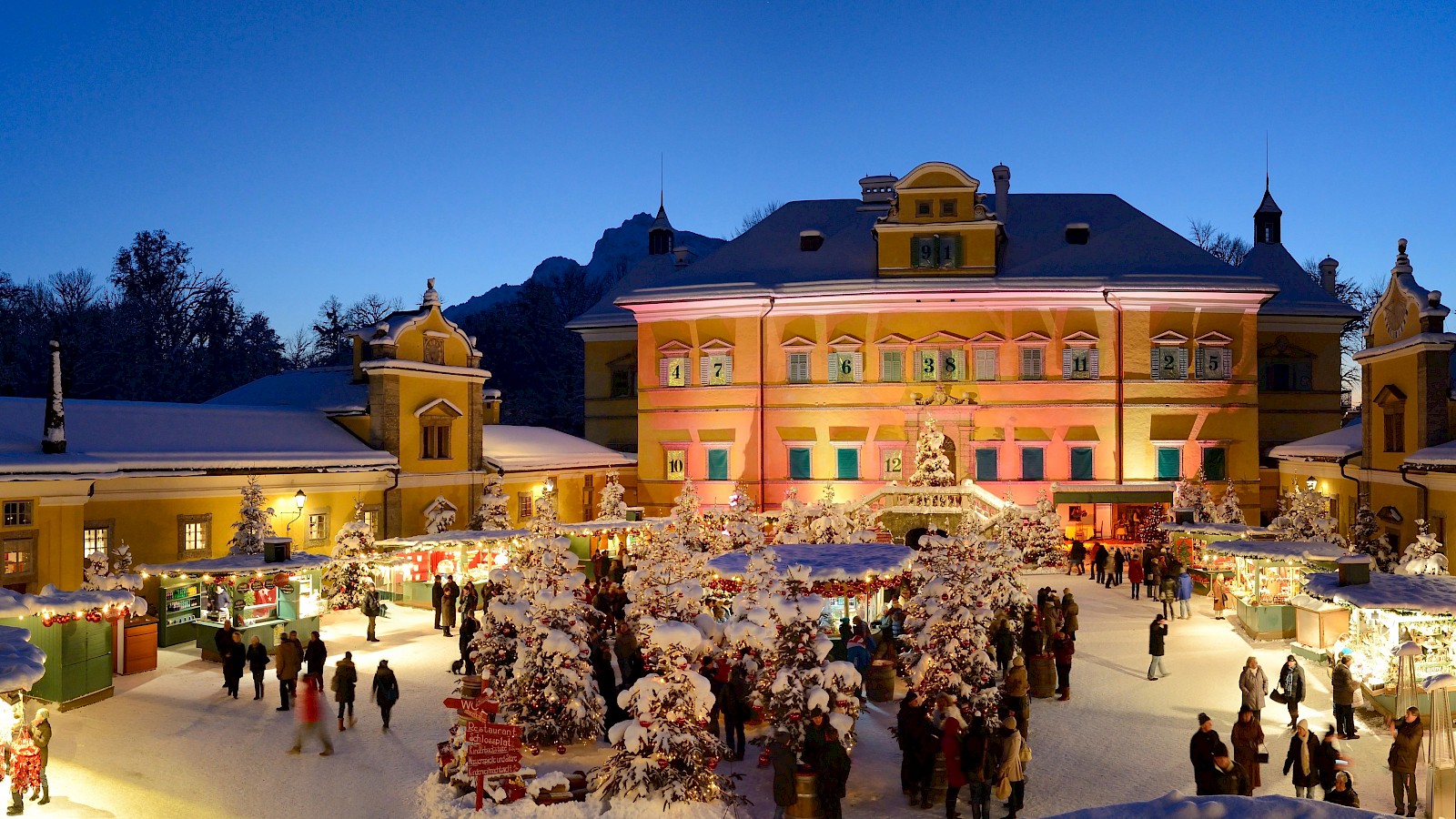 The festively decorated scene shows the atmospheric Christmas market in the courtyard of Hellbrunn Palace. In the center stand splendidly adorned Christmas trees, surrounded by numerous stalls illuminated with lights and typical wooden roofs. Many visitors stroll through the market, enjoying the pre-Christmas atmosphere. The palace in the background is bathed in magical violet light, completing the romantic image. The clear dusk enhances the effect of the lights and gives the place a fairytale ambiance. A charming example of European Advent traditions.