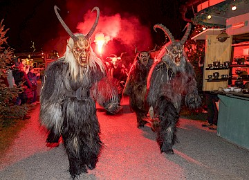 Eine furchteinflößende Krampusmaske mit wilden Zähnen, roten und schwarzen Farbtönen sowie zotteligem Fell dominiert das Bild. Der Hintergrund ist von Lichterketten erhellt, was auf ein vorweihnachtliches Fest hindeutet. Die Szene zeigt die traditionelle alpenländische Krampusfigur, die als düsterer Begleiter des Nikolaus auf Weihnachtsmärkten für Spannung sorgt.