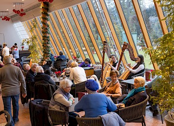 A snowy winter evening outside the Orangerie at Hellbrunn. Through the large windows, guests can be seen enjoying a festive atmosphere by candlelight. Outside, snow-covered trees are illuminated in magical pink light. The contrast between the cozy interior and the enchanted winter garden creates a fairytale-like, festive ambiance.