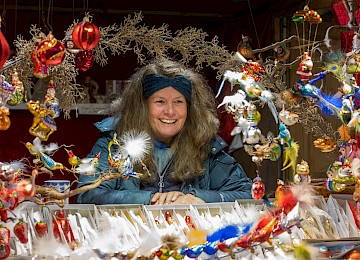 A blacksmith hammers a stamp onto a horseshoe on an anvil—creating a personalized good luck charm. The scene is warmly decorated with a lantern, fir branches, and festive lights, evoking traditional craftsmanship and a cozy Christmas market atmosphere.