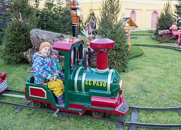 Ein Kind fährt begeistert mit einer kleinen, bunten Eisenbahn namens „El Paso“ durch eine verschneite Winterlandschaft. Umgeben von geschmückten Tannenbäumen und Weihnachtsdeko herrscht festliche Stimmung. Die Szene vermittelt kindliche Freude, Weihnachtszauber und familiäre Wärme auf einem stimmungsvollen Adventsmarkt.
